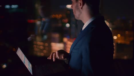 Late-at-Night-in-the-Office-Successful-Businessman-Holds-Laptop-while-Working-on-it.-In-the-Window-Business-District-View-with-City-Lights.