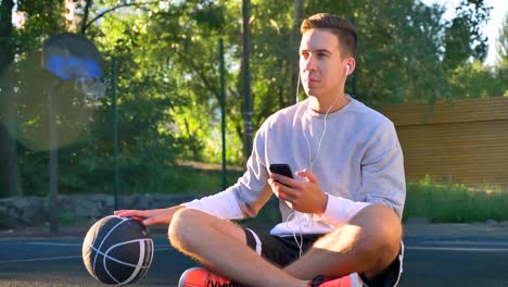 Young-man-sitting-on-basketball-court-and-dribbling-ball,-listening-music-through-earphones-and-holding-phone,-open-air-and-lens-flare