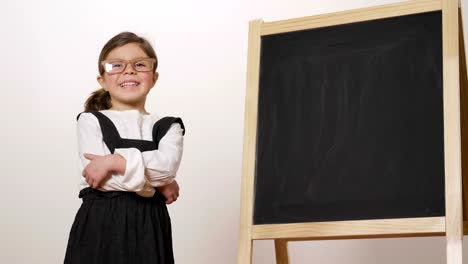 A-happy-girl-dressed-as-a-teacher-in-front-of-a-small-blackboard-holds-her-arms-folded-and-smiles.