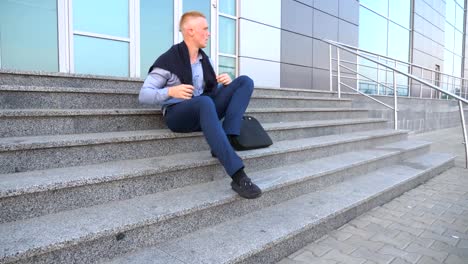 Dolly-shot-of-desperate-young-businessman-sitting-on-stairs-near-office-building-and-taking-off-cardigan-of-the-shoulders-with-anger-outdoor.-Upset-sad-manager-got-a-very-bad-news-and-crying.-Close-up