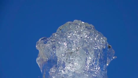 A-splash-of-clear-ice-water.-Closeup-of-light-splashing-water-in-sunny-fountain-in-park-in-summer-on-blue-sky-background.--Water-flow-of-fountain-fly-up-in-air-with-many-splashes.-Slow-motion.