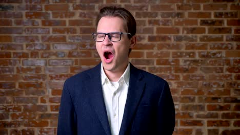 Yawning-caucasian-guy-is-standing-next-to-camera-on-brick-background-and-looking-tired-and-relaxed-indoor