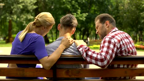 Caring-mother-and-dad-supporting-sad-teen-son-sitting-on-bench-in-park,-crisis