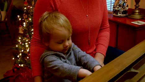 Caucasian-Toddler-Boy,-Sitting-at-a-Piano-with-Him-Mom,-Plays-on-the-Piano-with-a-decorated-Christmas-Tree-Behind-Them