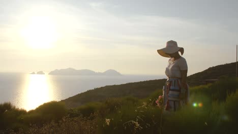 Beautiful-young-woman-wearing-fashion-colorful-dress-with-skirt-and-hat-carrying-flowers-in-basket-at-sunset-on-Ponza-Island-mountain-Italy.