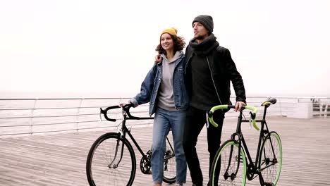 Modern-young-hipster-couple-walking-together-with-their-bikes-near-the-sea-at-autumn-day-embracing.-Walking-by-wooden-deck-in-daytime,-smiling-and-talking.-Slow-motion