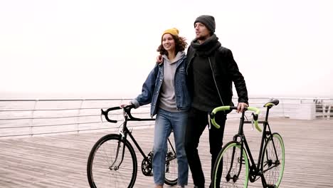 Pareja-moderna-joven-inconformista-caminando-junto-con-sus-bicicletas-cerca-del-mar-en-día-de-otoño-abrazando.-Caminando-por-cubierta-de-madera-durante-el-día,-sonriendo-y-hablando.-Cámara-lenta