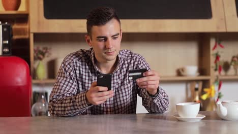 Handsome-man-making-purchase-online,-typing-credit-card-details-on-black-smartphone-in-the-modern-kitchen