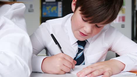 Estudiante-de-secundaria-de-hombre-usando-escritura-uniforme-en-ejercicio-libro-sentado-en-el-escritorio-en-el-aula