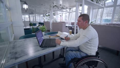 online-education,-successful-student-invalid-men-on-wheelchair-uses-modern-laptop-technology-to-learn-from-online-teaching-and-books-making-notes-in-notebook-close-up-sitting-at-table