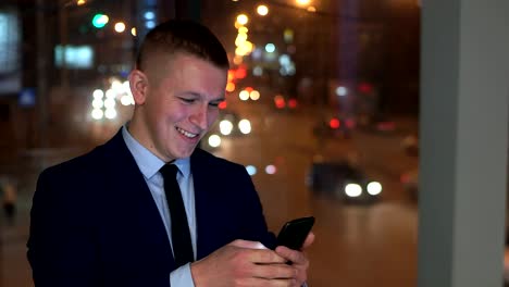 Der-Mann-Geschäftsmann-befasst-sich-mit-dem-Smartphone-glücklich,-glücklich.-Draußen-vor-dem-Fenster,-gegen-die-nächtliche-Stadt.