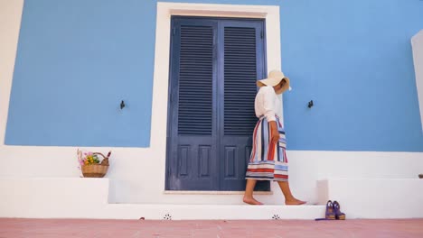 Mujer-joven-feliz-con-bicicleta-caminando-frente-a-patio-de-la-puerta-de-la-casa-azul.-Camisa-de-moda-blanco,-gran-sombrero,-falda-colorida-y-gafas-de-sol.-Isla-de-Ponza,-Italia.