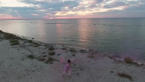 En-vista-de-sensual-joven-con-maquillaje-brillante-en-un-color-de-rosa-Vestido-de-baile-con-bombas-de-humo-en-la-orilla-del-río.-El-baile-de-una-chica-muy-dulce.-Cámara-lenta.-El-rodaje-de-los-aviones-no-tripulados.