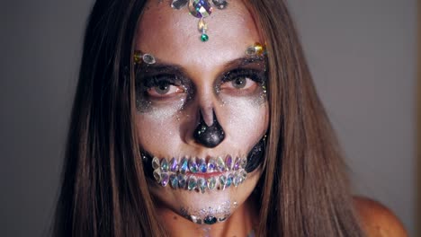 Young-girl-with-creative-halloween-face-art-in-the-studio-with-grey-background.-Portrait-of-glamorous-skull-with-rhinestones-and-sequins.-Professional-make-up-for-the-celebration.-Slow-motion