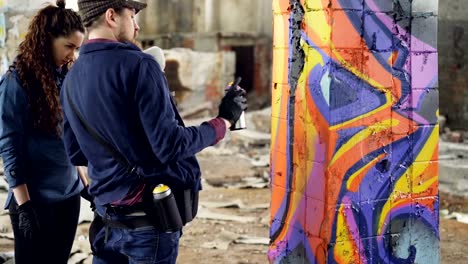 Urban-artist-handsome-bearded-man-is-teaching-amateur-student-to-work-with-aerosol-paint-while-decorating-old-industrial-building-with-abstract-graffiti.