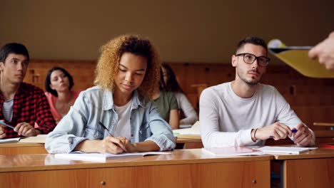 Smart-guy-in-glasses-student-is-talking-to-teacher-sitting-at-desk-in-lecture-hall-with-young-people-around-him.-Higher-education,-youth-and-getting-knowledge-concept.