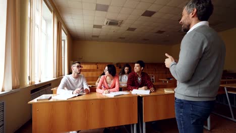 Kleine-Gruppe-von-Studenten-ist-im-Gespräch-mit-Lehrer,-junger-Mann-hob-Hand-und-Frage,-lächelnden-Erzieher-hört-zu-und-gestikulierte.-Lern--und-Lehrkonzept.