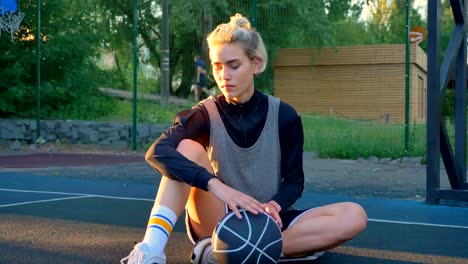 Beautiful-blonde-female-basketball-player-sitting-on-ground-and-playing-with-ball,-park-in-background