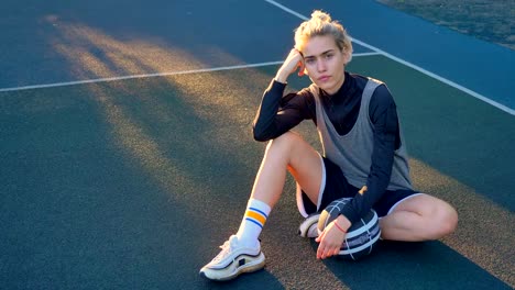 Pensive-charming-young-female-basketball-player-sitting-on-ground-and-looking-at-camera,-holding-ball,-outdoors