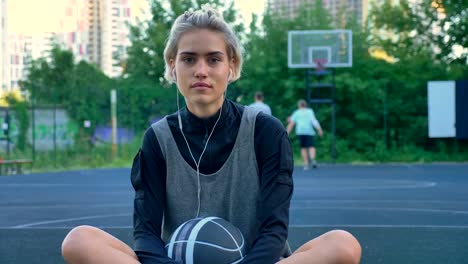 Lächelnden-jungen-weiblichen-Basketball-Player-Musik-hören-über-Kopfhörer-und-sitzen-auf-dem-Hof,-Männer-spielen-auf-Hintergrund-im-park