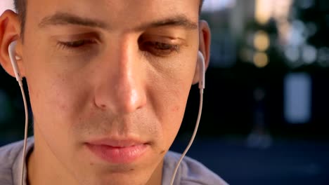 Seria-guapo-joven-escuchar-música-mediante-auriculares-y-mirando-a-cámara,-gente-jugando-al-baloncesto-en-fondo