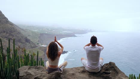 Un-hombre-y-una-mujer-sentada-en-la-cima-de-una-montaña-mirando-el-mar-sentado-en-una-piedra-meditando-en-posición-de-loto.-La-vista-desde-la-parte-posterior.-Islas-Canarias