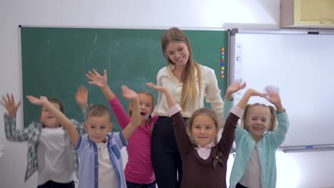 Escuela-primaria,-grupo-de-divertidos-niños-saltando-y-agitando-las-manos-cerca-de-la-maestra-en-el-fondo-de-la-Junta