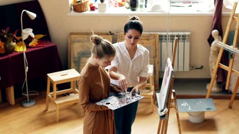 Gut-aussehende-junge-Frau-hält-Farben-und-Malerei-Blumen-unter-Anleitung-hilfreich-Lehrers-im-Arbeitsraum-der-Kunstschule-zusammen-stehen-und-reden.