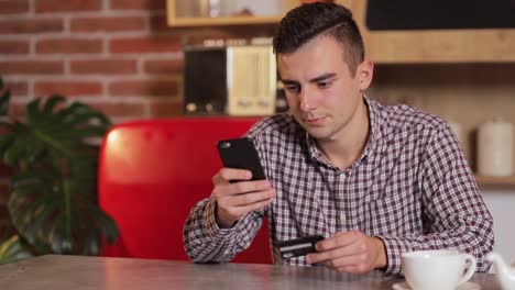 Handsome-man-making-purchase-online,-typing-credit-card-details-on-black-smartphone-in-the-modern-kitchen