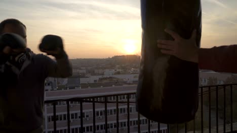 Hands-of-trainer-holds-boxing-bag-while-man-boxer-punching-on-training-in-sunset,-sportsman-practicing,-power-training,-strong-guy-hard-exercising,-strength-exercises,-workout,-handheld,-sunny-day.