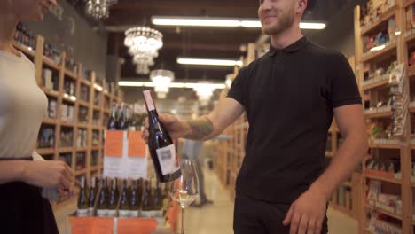 Cute-short-haired-girl-seller-offers-wine-to-the-visitor.-Male-buyer-takes-a-bottle-of-wine-offered-by-the-seller-and-smiles-happily.-Bearded-guy-with-tattoos-on-his-arm-wants-to-buy-good-wine-at-a-liquor-store.