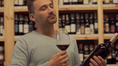 Portrait-of-handsome-male-who-smelling-wine-glass.-A-guy-with-a-bottle-of-wine-and-a-wine-glass-in-his-hands-standing-on-the-background-of-shelves-with-alcoholic-beverages.