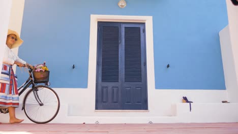 Mujer-joven-feliz-con-bicicleta-caminando-frente-a-patio-de-la-puerta-de-la-casa-azul.-Camisa-de-moda-blanco,-gran-sombrero,-falda-colorida-y-gafas-de-sol.-Isla-de-Ponza,-Italia.