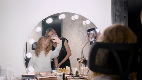 female-client-of-beauty-shop-is-taking-selfie-with-professional-makeup-artist-in-front-of-mirror-with-lamps