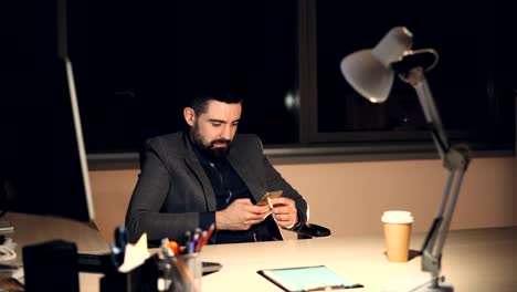 Atractivo-joven-barbudo-está-utilizando-smartphone-pantalla-táctil-y-sonriendo-trabajando-en-la-oficina-por-la-noche-solo.-Comunicación-y-concepto-de-la-tecnología-moderna.