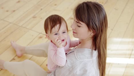 Über-dem-Blick-auf-die-junge-Mutter,-die-zu-Hause-auf-dem-Hartholzboden-sitzt-und-ihre-entzückende-Babytochter-einlullen,-streicheln-und-küssen