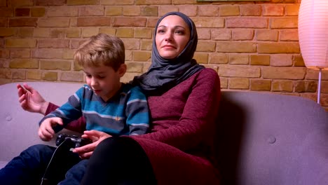 Konzentrierter-kleiner-Junge-und-seine-muslimische-Mutter-im-Hijab-spielen-Videospiel-mit-Joystick-zusammen-zu-Hause.