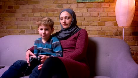 Konzentrierter-kleiner-Junge,-der-Videospiel-spielt,-und-seine-muslimische-Mutter-im-Hijab-versucht,-Joystick-zu-holen,-um-zu-versuchen,-zu-spielen.