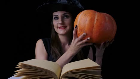 Attractive-witch-on-a-black-background-in-front-of-an-open-spell-book,-peeks-out-from-behind-a-pumpkin-and-smiles-every-time-her-mood-changes,-halloween-concept.