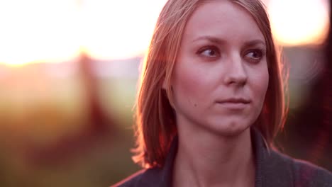 Portrait-of-young-pensive-woman-at-the-park-in-the-rays-of-the-setting-sun,-looking-into-the-distance,-close-up,-slow-mo