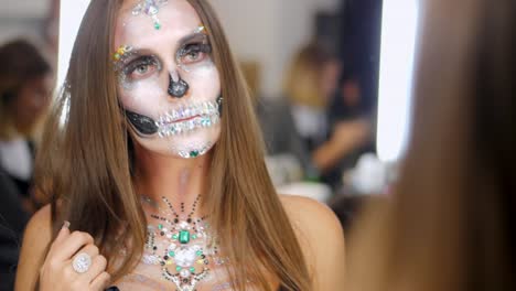 Chica-joven-con-arte-creativo-de-la-cara-de-halloween-mirando-en-el-reflejo-del-espejo-en-el-vestidor.-Retrato-de-glamour-cráneo-con-pedrería-y-lentejuelas.-Maquillaje-profesional-para-la-celebración