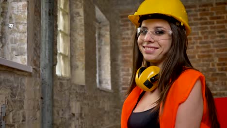 Joven-encantadora-atractivas-mujeres-en-casco,-auriculares-y-gafas-mirando,-señalando-en-la-cámara,-sonriendo,-mujer-builder,-edificio-de-ladrillo-de-fondo