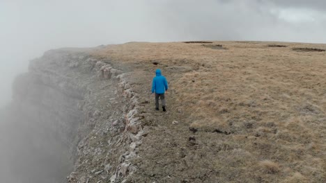 Hiker-man-in-a-blue-jacket-with-backpack-is-walking-and-standing-on-the-steep-edge-of-mountain-plateau-above-the-clouds.-Aerial-view.-Drone-is-orbiting