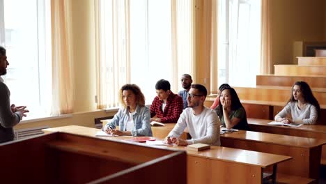 Smart-guy-student-is-raising-hand-and-talking-to-professor-while-fellow-students-are-listening-to-them-and-smiling.-Pupil-and-teacher-relations-concept.