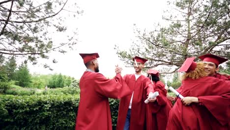 Slow-motion-of-friends-graduating-students-doing-high-five-and-embracing-each-other-wearing-mortarboard-caps-and-gowns-and-holding-diplomae.-Campus,-events-and-people-concept.