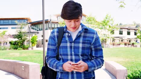 Male-High-School-Student-Checking-Messages-On-Mobile-Phone-Outside-College-Buildings