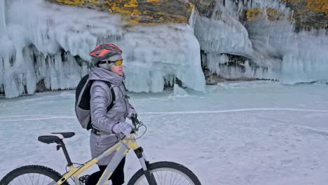 Woman-is-walking-beside-bicycle-near-the-ice-grotto.-The-rock-with-ice-caves-and-icicles-is-very-beautiful.-The-girl-is-dressed-in-silvery-down-jacket,-backpack-and-helmet.-The-traveler-is-ride-cycle.