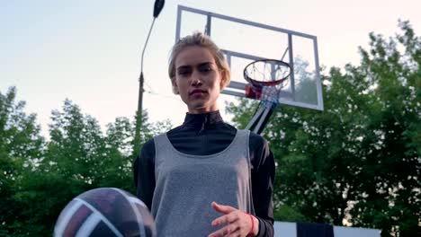 Jugador-de-baloncesto-femenina-rubia-atractiva-jugando-con-la-pelota-y-mirando-a-cámara,-en-el-parque,-durante-el-día