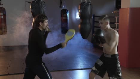 A-professional-boxer-with-his-coach-in-the-ring-trains-the-speed-of-strikes-and-reactions.-The-man-in-the-gym-is-training-against-the-backdrop-of-boxing-pears,-above-the-floor-a-dull-gray-haze