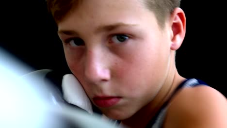 The-boxer-boy-stands-on-a-dark-background,-the-boy-has-a-gray-T-shirt,-his-face-is-almost-covered-in-gloved-hands.-The-boy-has-light-short-hair-and-dark-eyes